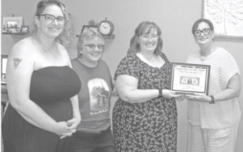 Sara Comer accepts the “first dollar” plaque from Chamber of Commerce representative Rebecca Spooner (far right).