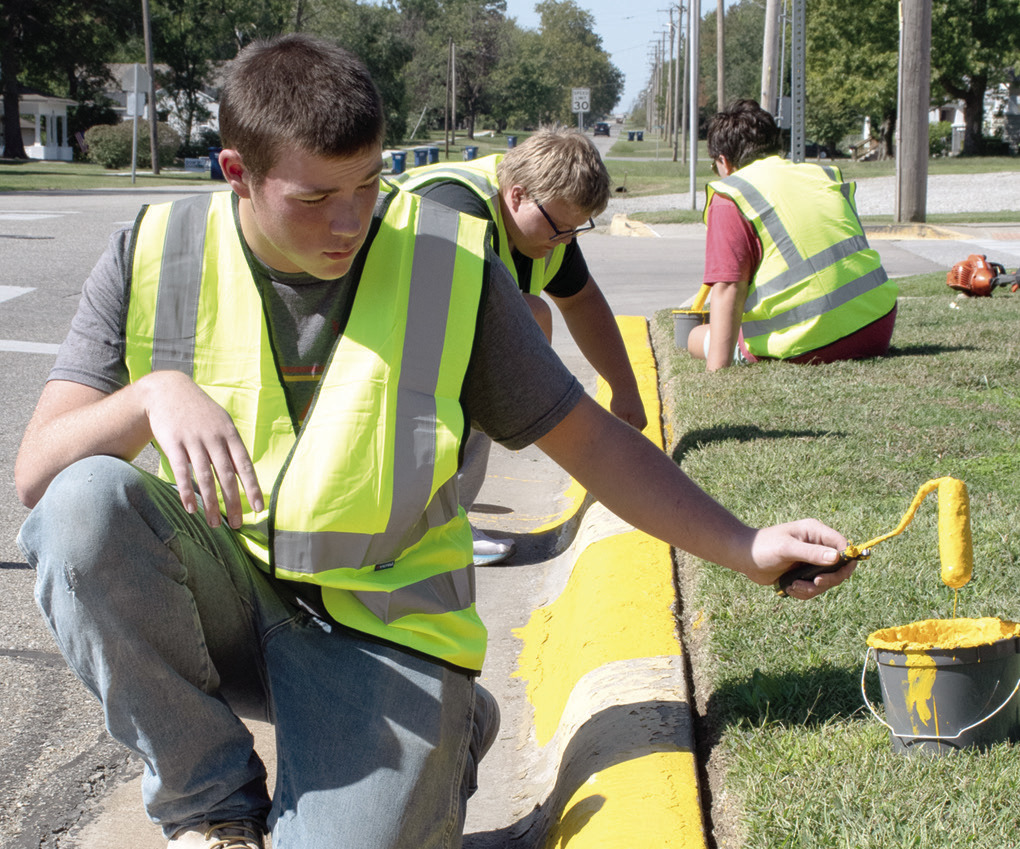 Above senior Jackson Wolken assisted with painting city curbs as part of the Community Day of Service held Wednesday afternoon. Junior Jayden Pease and senior Asher Lloyd also assisted. At right drivers downtown Thursday were greeted with this sign on Maple Street as crews with Joplin Striping worked to complete painting of downtown parking spaces and road striping ahead of the annual Columbus Day Festival.