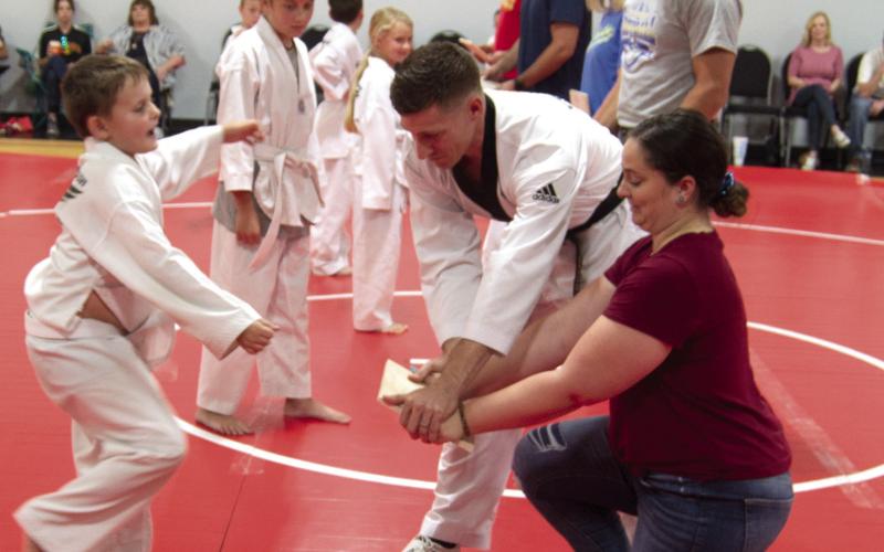 Student Jaik Hayter kicks at the board held by his mother Lauren Vaughn and Master Vigil. Other students and parents look on during the demonstration.