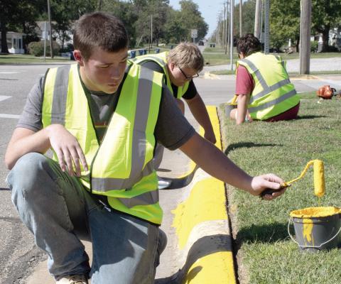 Above senior Jackson Wolken assisted with painting city curbs as part of the Community Day of Service held Wednesday afternoon. Junior Jayden Pease and senior Asher Lloyd also assisted. At right drivers downtown Thursday were greeted with this sign on Maple Street as crews with Joplin Striping worked to complete painting of downtown parking spaces and road striping ahead of the annual Columbus Day Festival.