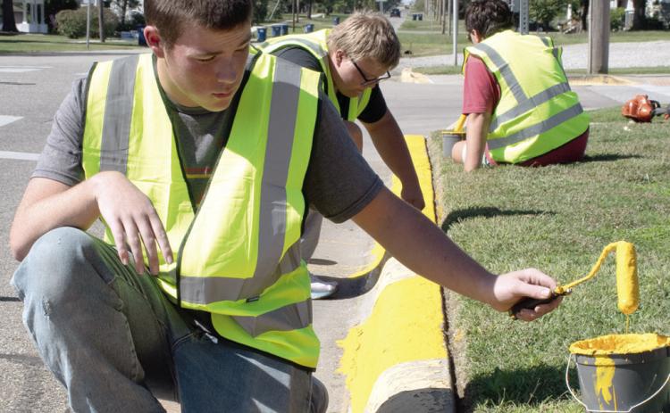Above senior Jackson Wolken assisted with painting city curbs as part of the Community Day of Service held Wednesday afternoon. Junior Jayden Pease and senior Asher Lloyd also assisted. At right drivers downtown Thursday were greeted with this sign on Maple Street as crews with Joplin Striping worked to complete painting of downtown parking spaces and road striping ahead of the annual Columbus Day Festival.