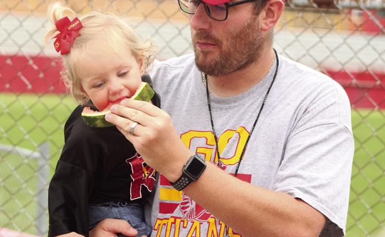 Titan Assistant Coach Matt Crain and daughter Sloan enjoy watermelon served following the Annual Columbus Lawn Chair Scrimmage, Saturday morning at Titan Stadium. Watermelon was served by Clint Saporito.