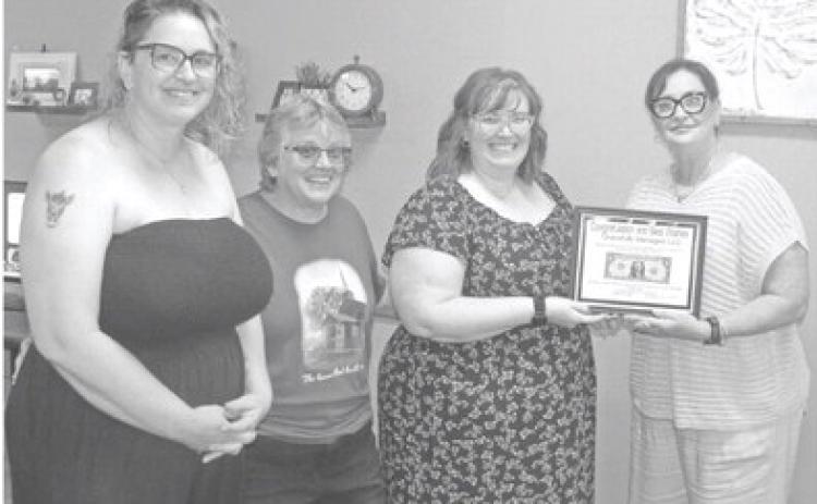 Sara Comer accepts the “first dollar” plaque from Chamber of Commerce representative Rebecca Spooner (far right).